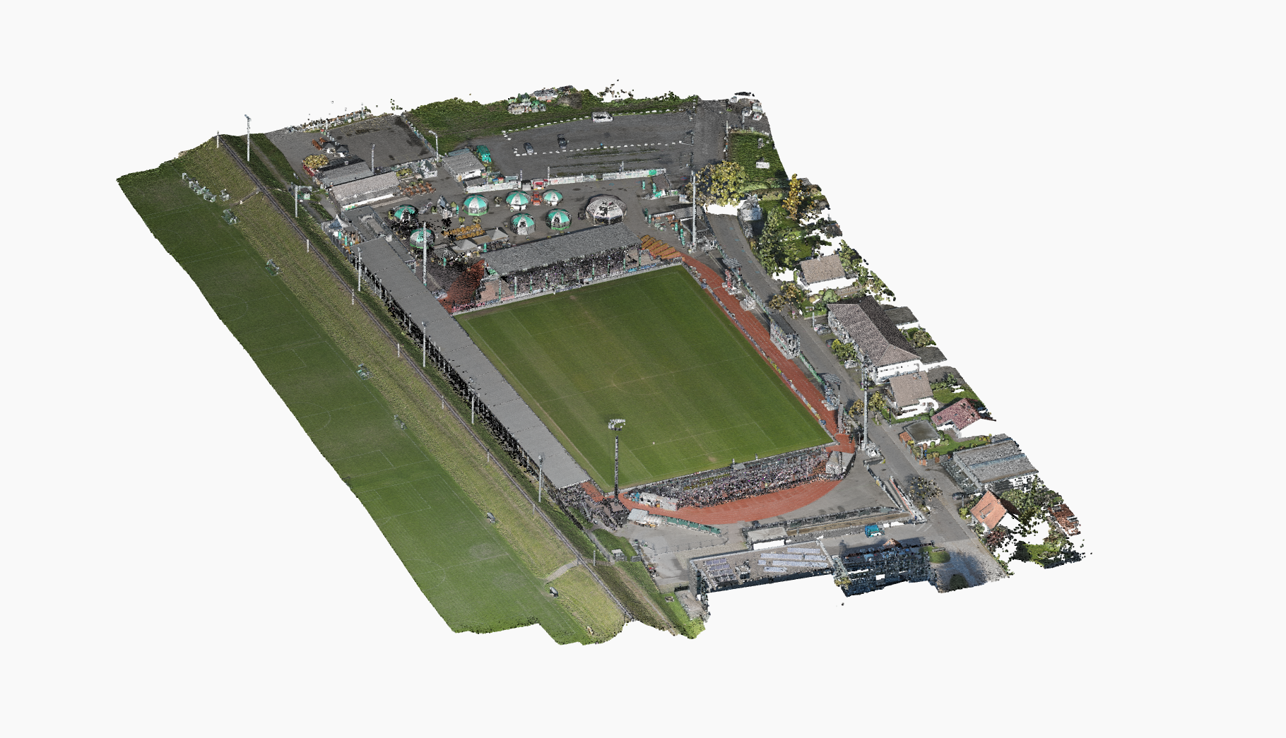 Rendu aérien en 3D d'un stade de football entouré de bâtiments, de routes et de champs verts, avec une foule visible dans les gradins et un match se déroulant sur le terrain.