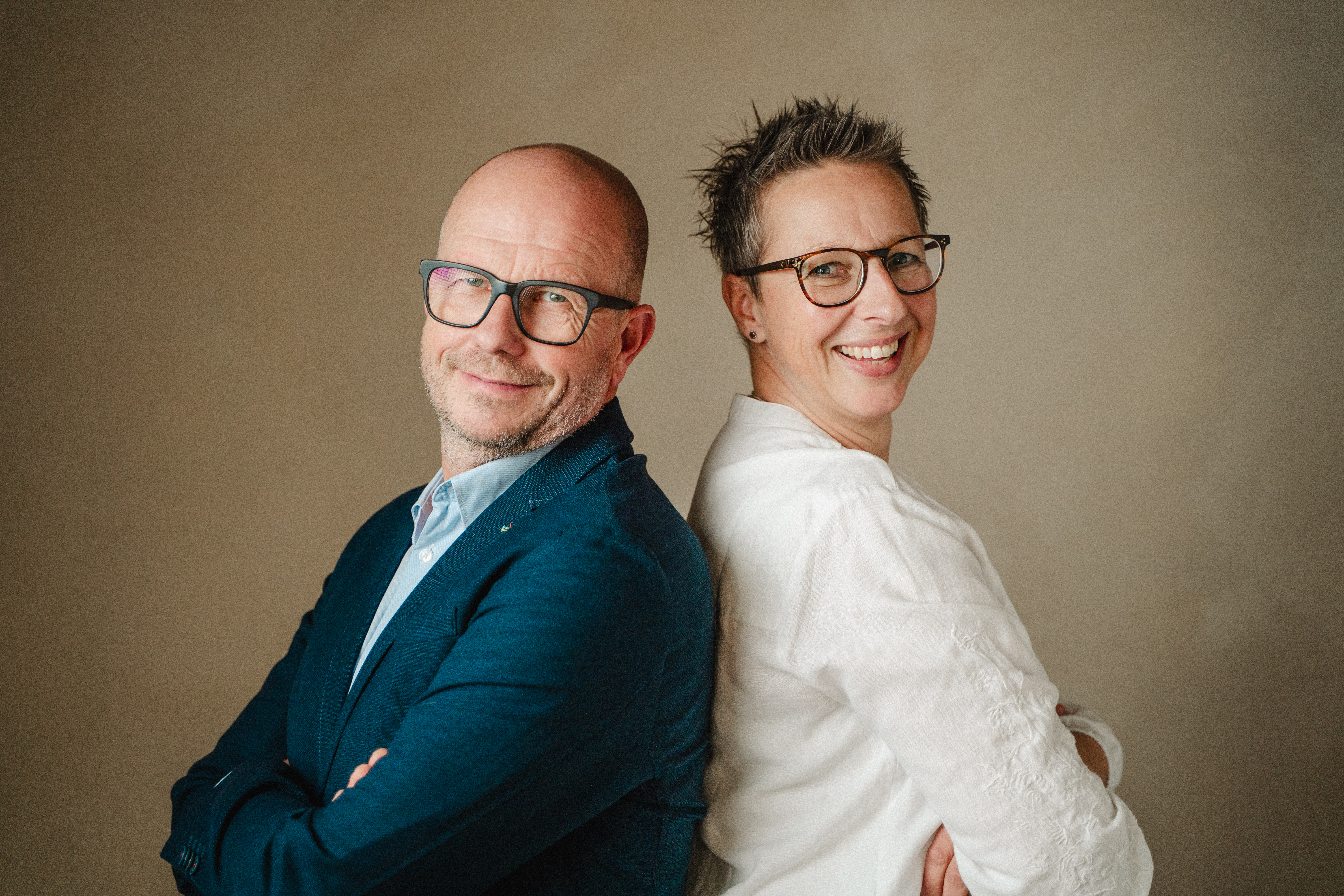 Ein Mann und eine Frau stehen Rücken an Rücken, beide lächeln in die Kamera. Der Mann trägt eine Brille, einen blauen Blazer und ein hellblaues Hemd. Die Frau hat kurze Haare, eine Brille und ein weißes Hemd. Neutraler beiger Hintergrund.