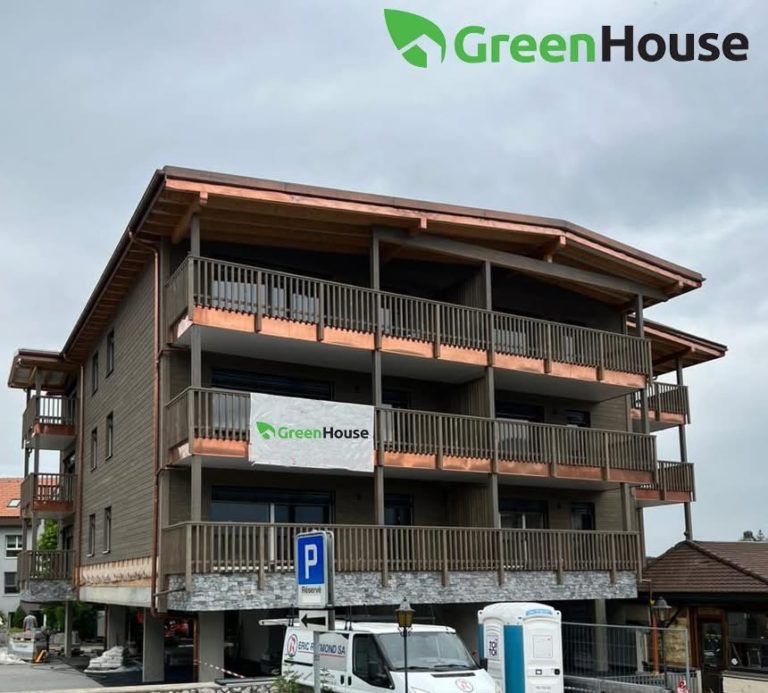 Ein dreistöckiges Holzgebäude mit Balkonen, an dessen Fassade ein GreenHouse-Banner hängt. Das GreenHouse-Logo erscheint in der oberen rechten Ecke. Auf der Straße darunter sind ein blaues Parkschild und Autos zu sehen.
