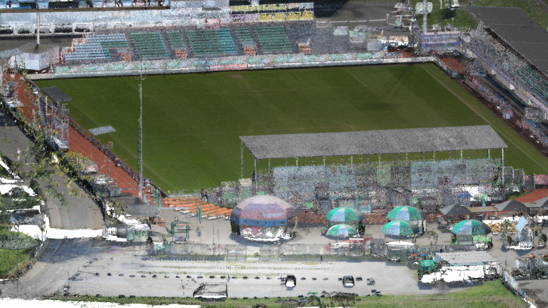 Vue aérienne d'un stade de football avec des tribunes vertes, un terrain en herbe et plusieurs tentes et véhicules installés à l'extérieur de la zone d'entrée.