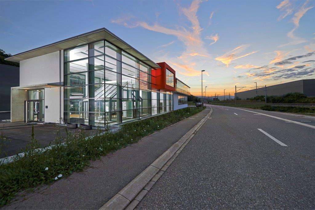Un bâtiment moderne en verre avec un accent rouge surplombe une route déserte au coucher du soleil, avec des nuages spectaculaires et une lumière douce dans le ciel. Des mauvaises herbes poussent le long du trottoir à côté du bâtiment.