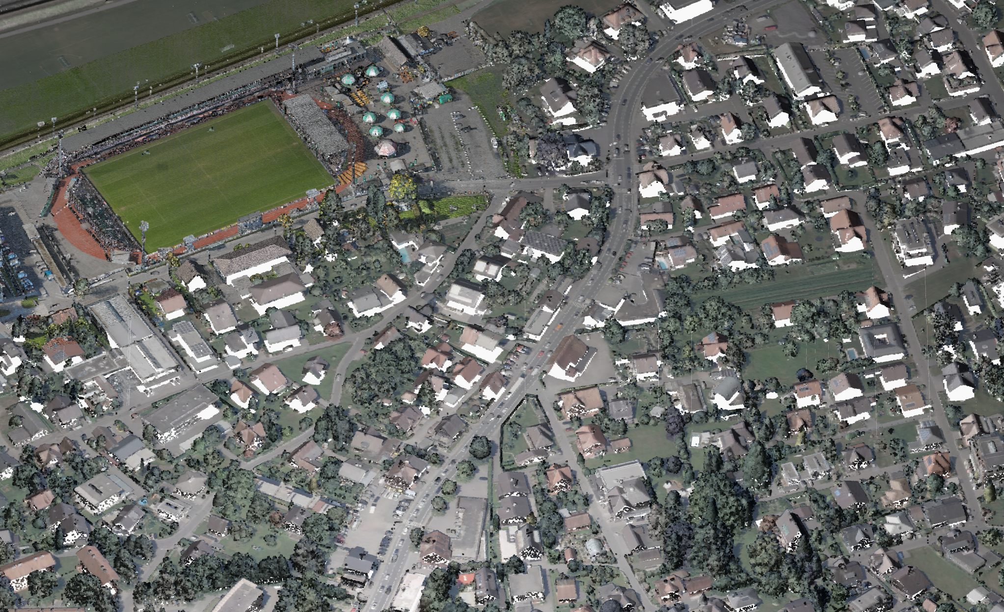 Vue aérienne d'un quartier de banlieue avec de nombreuses maisons, de la verdure et un grand stade de sport rempli de monde sur le côté gauche de l'image. Des routes traversent la zone résidentielle.