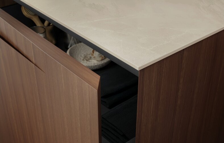 Gros plan sur une armoire en bois partiellement ouverte, contenant des serviettes noires pliées et un bol blanc texturé posé sur une étagère sous un plan de travail en marbre clair.