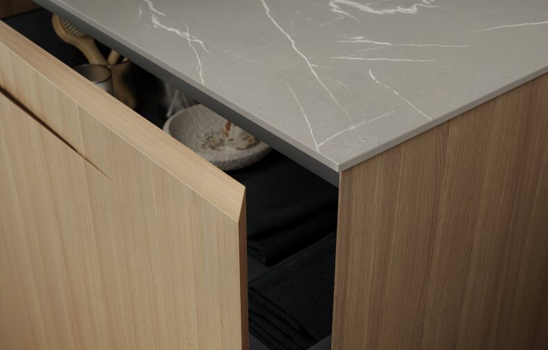 Une porte en bois partiellement ouverte laisse entrevoir des serviettes noires pliées et un bol à motifs blancs posé sous un plan de travail en marbre gris avec des veines blanches.
