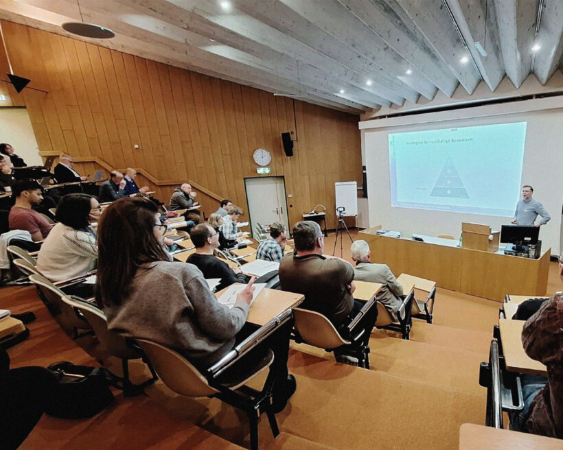 Eine Gruppe von Personen sitzt in einem Hörsaal auf gestuften Sitzen und hört einem Vortragenden zu, der vor einem Projektor steht, auf dem eine Folie mit einem Pyramidendiagramm zu sehen ist. Der Raum hat Holzwände und eine hohe Decke.