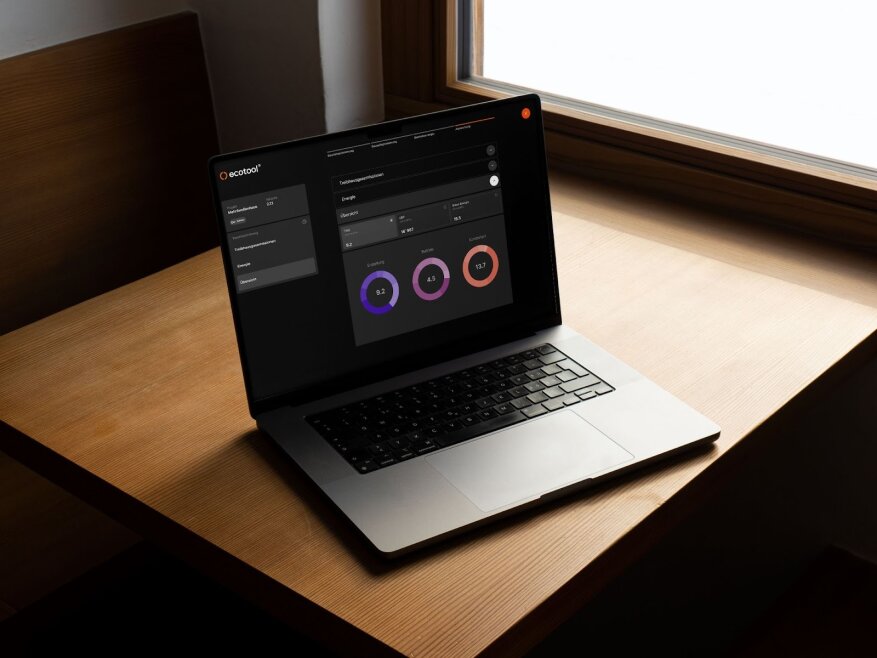 Ein Laptop auf einem Holztisch in der Nähe eines Fensters zeigt ein dunkles Armaturenbrett mit Diagrammen und Grafiken, darunter zwei kreisförmige Fortschrittsanzeigen und Navigationsmenüs auf der linken Seite des Bildschirms.