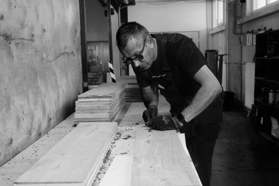 Ein Mann mit Brille und Handschuhen bearbeitet ein Stück Holz an einer Werkbank in einer Schreinerei. Um ihn herum sind in dem gut beleuchteten Raum Stapel von Holzbrettern und Werkzeugen zu sehen.