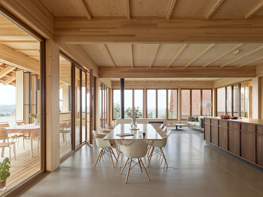 Ein ger&auml;umiger, moderner Ess- und Wohnbereich mit Holzdecken und gro&szlig;en Fenstern, mit wei&szlig;en St&uuml;hlen um einen Tisch, offenen Regalen und viel nat&uuml;rlichem Licht mit Blick auf eine Au&szlig;enterrasse und eine malerische Aussicht.