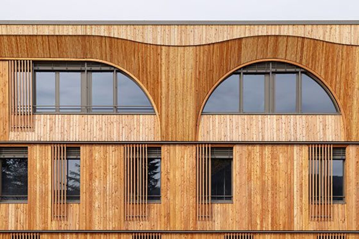 L'image montre une façade de bâtiment moderne faite de lamelles de bois verticales, avec de grandes fenêtres arquées à l'étage supérieur et des fenêtres rectangulaires avec des accents en bois en dessous.