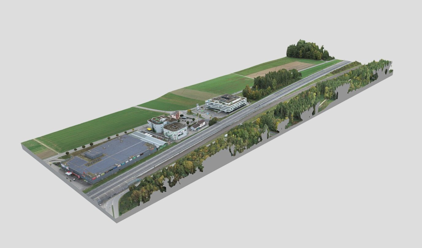 Rendu aérien en 3D d'une zone rurale comprenant des routes, des parkings, plusieurs bâtiments, des champs et des groupes d'arbres, avec une autoroute parallèle aux structures et au paysage.