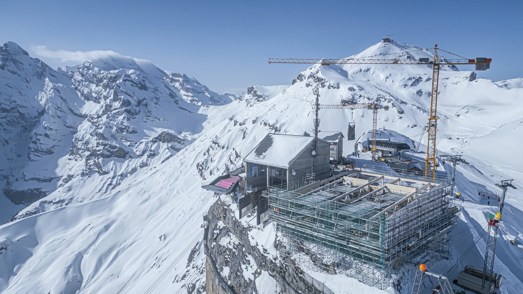 Un chantier minier avec des grues et des échafaudages se trouve sur un sommet enneigé, entouré de montagnes escarpées et enneigées sous un ciel bleu clair.