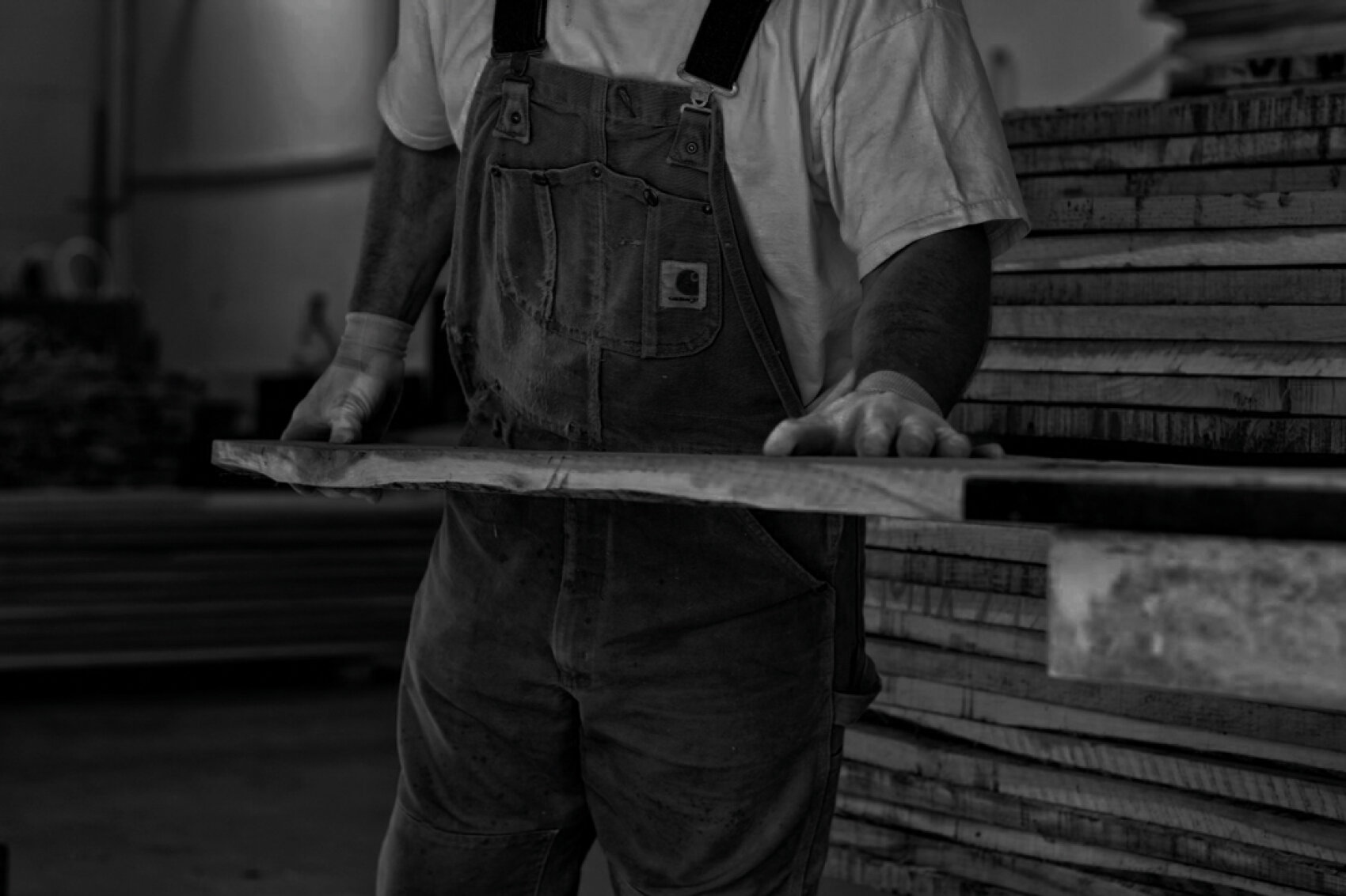 Une personne portant une combinaison et un T-shirt vérifie ou travaille une planche de bois dans un atelier, avec des piles de bois en arrière-plan. L'image est en noir et blanc.