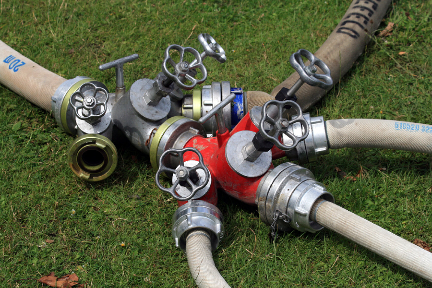 Ein Hydrantenanschluss mit mehreren Schläuchen, der auf grünem Gras liegt. Die Metallventile und -anschlüsse sind sichtbar, wobei zwei Schläuche mit einem roten Abschnitt und andere mit silbernen Abschnitten verbunden sind.
