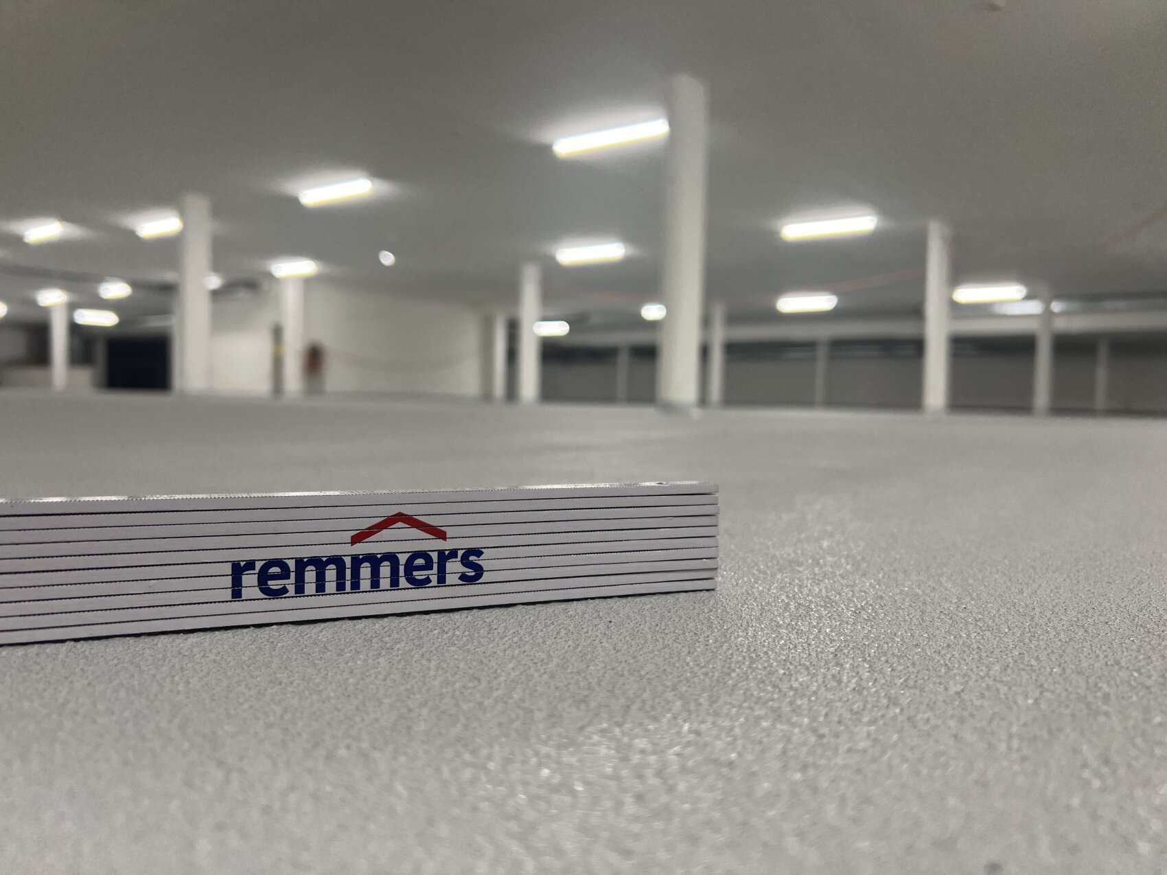 Nahaufnahme eines gefalteten Lineals mit dem Remmers-Logo auf einem grauen, strukturierten Boden in einem hell erleuchteten, geräumigen, leeren Innenraum mit weißen Säulen und Deckenleuchten.