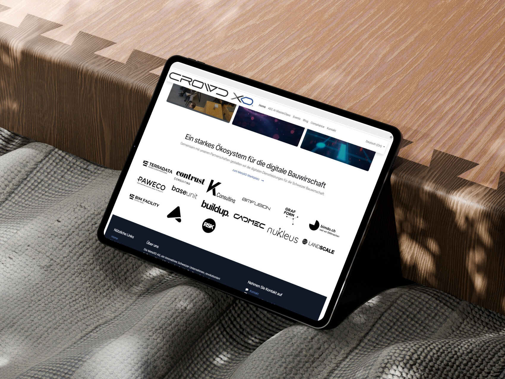 Une tablette sur laquelle s'affiche un site web avec les logos de différentes entreprises est posée sur un plafond gris texturé et une table en bois avec des motifs de bordures en zigzag.