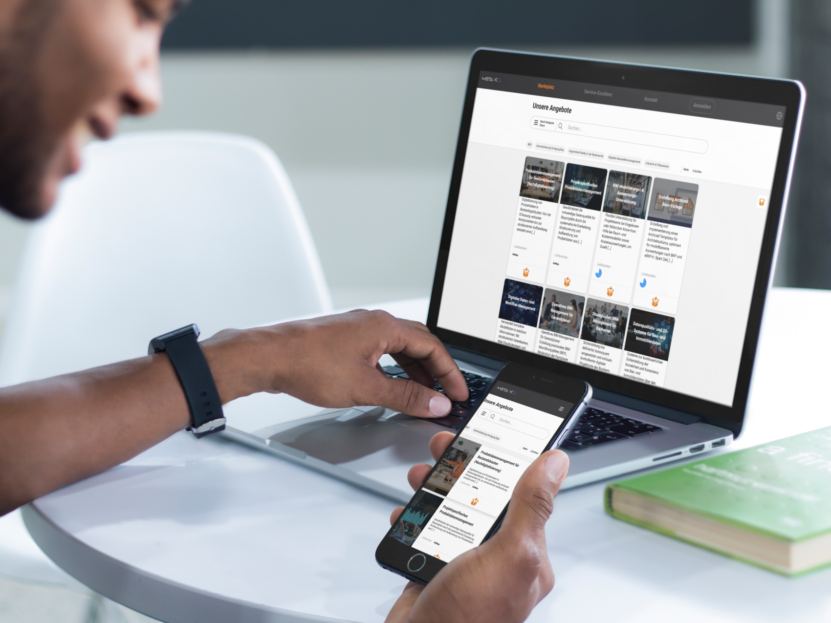Une personne est assise à une table et utilise un smartphone et un ordinateur portable, qui affichent tous deux le même site web avec une grille d'images et de texte. Un livre vert fermé et une chaise blanche sont également posés sur la table.