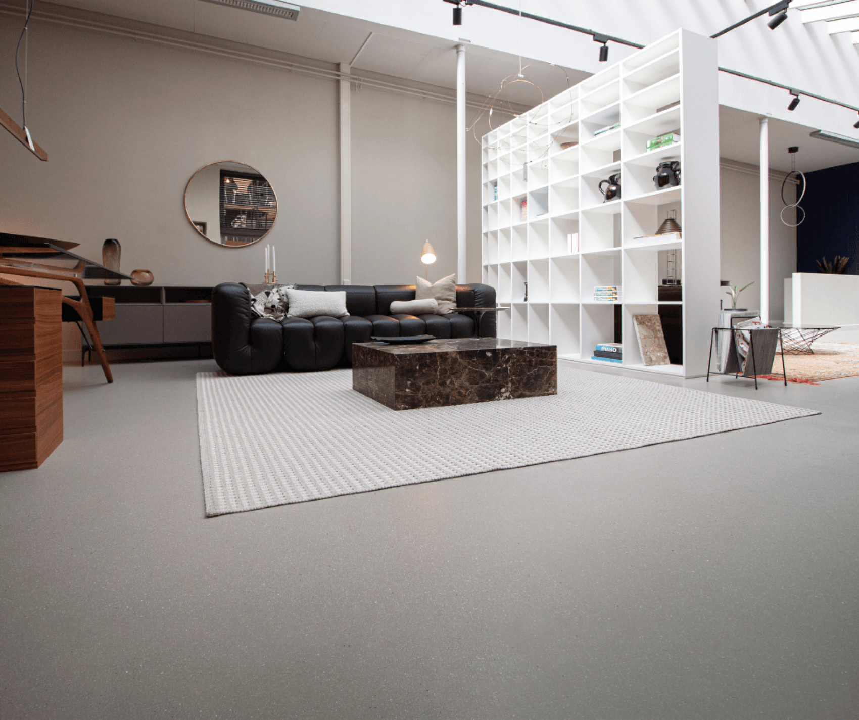 Ein modernes Wohnzimmer mit einem schwarzen Sofa, einem Couchtisch aus Marmor, einem weißen Teppich und offenen weißen Regalen, die den Raum unterteilen. Der Raum ist minimalistisch eingerichtet und hat hohe Decken mit Oberlichtern.